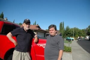 Two adult men standing beside a red pickup truck on a quiet residential street, representing a father and son having a difficult conversation about aging and future care