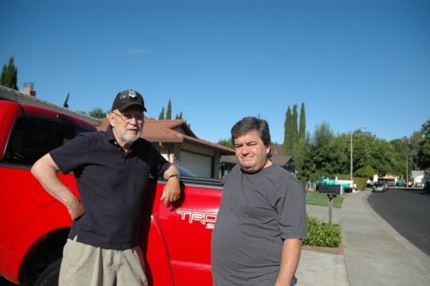 Two adult men standing beside a red pickup truck on a quiet residential street, representing a father and son having a difficult conversation about aging and future care